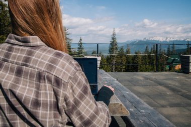 Genç, güzel, ince, dalgalı saçlı, ekose gömlekli bir kız eski ahşap bir masada oturuyor ve bir dizüstü bilgisayarın başında dağların arka planına karşı çalışıyor. Arkadan bak..