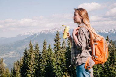 Ekose gömlekli, bol saçlı, turuncu sırt çantalı genç bir kız Karpatlar 'daki dağların arka planında parlak bir termal bardaktan çay içiyor. Arkadan görünüm.