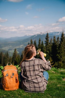 A young, slender girl with loose hair in a plaid shirt and jeans with an orange backpack drinks tea from a thermal mug sitting on the green grass against the backdrop of the mountains.