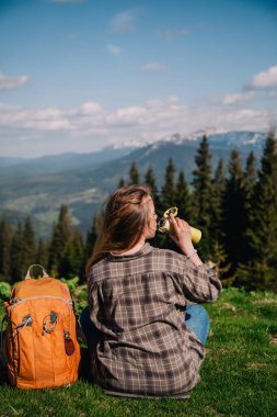 A young, slender girl with loose hair in a plaid shirt and jeans with an orange backpack drinks tea from a thermal mug sitting on the green grass against the backdrop of the mountains.