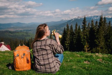 A young, slender girl with loose hair in a plaid shirt and jeans with an orange backpack drinks tea from a thermal mug sitting on the green grass against the backdrop of the mountains.