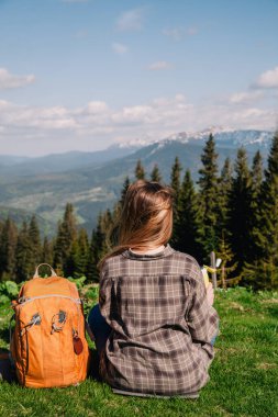 A young, slender girl with loose hair in a plaid shirt and jeans with an orange backpack drinks tea from a thermal mug sitting on the green grass against the backdrop of the mountains.