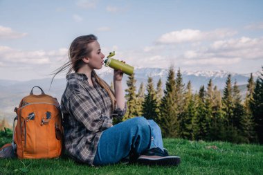 Genç, ince saçlı, ekose gömlekli, turuncu sırt çantalı, kot pantolonlu bir kız, dağların arka planında yeşil çimlerin üzerinde oturan termal bardaktan çay içer..