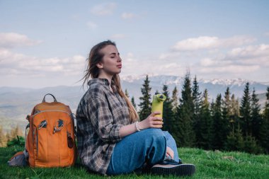 Genç, ince saçlı, ekose gömlekli, turuncu sırt çantalı, kot pantolonlu bir kız, dağların arka planında yeşil çimlerin üzerinde oturan termal bardaktan çay içer..