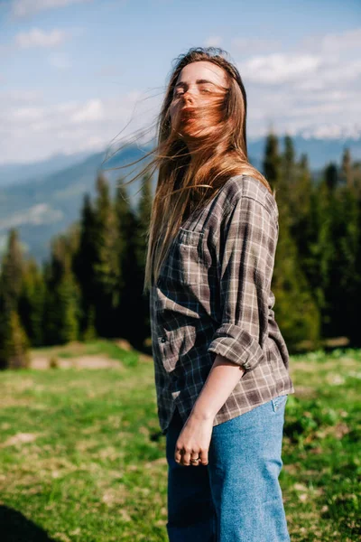 Une jeune fille mince avec les cheveux lâches dans une chemise à carreaux et un jean pose pour le temps ensoleillé dans les montagnes — Photo