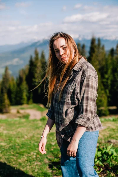 Une jeune fille mince avec les cheveux lâches dans une chemise à carreaux et un jean pose pour le temps ensoleillé dans les montagnes — Photo