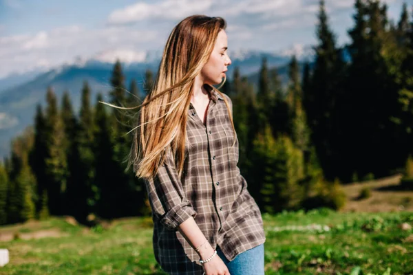 Genç, ince saçlı, ekose gömlekli ve kot pantolonlu bir kız dağlarda güneşli hava için poz veriyor.