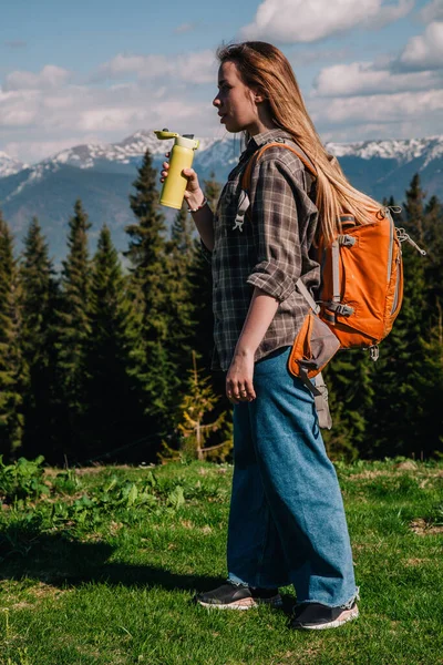 A young, girl in a plaid shirt and jeans with loose hair with an orange backpack drinks tea from a bright thermal cup against the backdrop of the mountains in the Carpathians. view from the back. — Stock Photo, Image