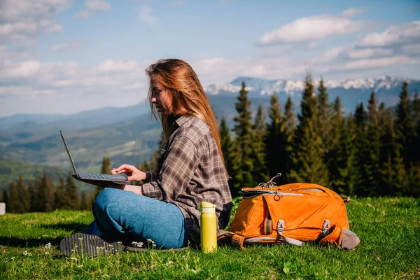 Ekose gömlek ve turuncu sırt çantalı bol saçlı genç, narin bir kız, yeşil çimlerin üzerinde, dağların arka planında oturuyor..