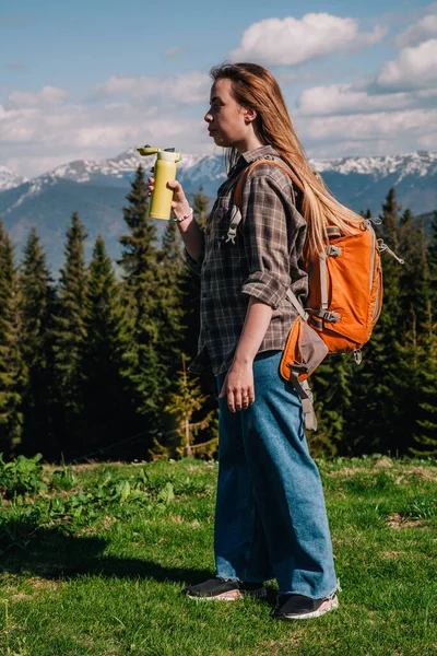 A young, girl in a plaid shirt and jeans with loose hair with an orange backpack drinks tea from a bright thermal cup against the backdrop of the mountains in the Carpathians. view from the back. —  Fotos de Stock