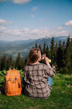 A young, slender girl with loose hair in a plaid shirt and jeans with an orange backpack drinks tea from a thermal mug sitting on the green grass against the backdrop of the mountains.