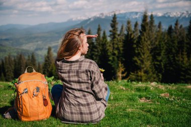 A young, slender girl with loose hair in a plaid shirt and jeans with an orange backpack drinks tea from a thermal mug sitting on the green grass against the backdrop of the mountains.