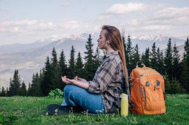 Ekose gömlekli, kot pantolonlu, turuncu sırt çantalı genç, narin bir kız yeşil çimlerin üzerinde, dağların arka planında meditasyon yapıyor..