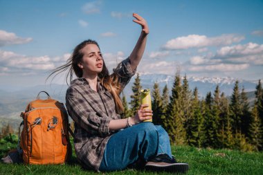 Genç, ince saçlı, ekose gömlekli, turuncu sırt çantalı, kot pantolonlu bir kız, dağların arka planında yeşil çimlerin üzerinde oturan termal bardaktan çay içer..