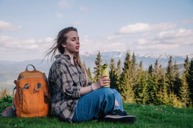 Genç, ince saçlı, ekose gömlekli, turuncu sırt çantalı, kot pantolonlu bir kız, dağların arka planında yeşil çimlerin üzerinde oturan termal bardaktan çay içer..