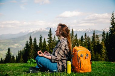 A young, slender girl in a plaid shirt and jeans with an orange backpack meditates sitting on the green grass against the backdrop of mountains.