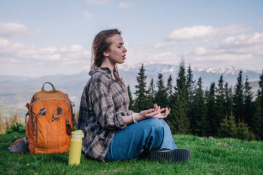 Ekose gömlekli, kot pantolonlu, turuncu sırt çantalı genç, narin bir kız yeşil çimlerin üzerinde, dağların arka planında meditasyon yapıyor..