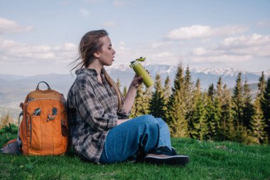 Genç, ince saçlı, ekose gömlekli, turuncu sırt çantalı, kot pantolonlu bir kız, dağların arka planında yeşil çimlerin üzerinde oturan termal bardaktan çay içer..