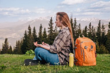 Ekose gömlekli, kot pantolonlu, turuncu sırt çantalı genç, narin bir kız yeşil çimlerin üzerinde, dağların arka planında meditasyon yapıyor..