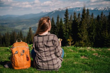 A young, slender girl with loose hair in a plaid shirt and jeans with an orange backpack drinks tea from a thermal mug sitting on the green grass against the backdrop of the mountains.