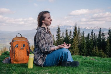 Ekose gömlekli, kot pantolonlu, turuncu sırt çantalı genç, narin bir kız yeşil çimlerin üzerinde, dağların arka planında meditasyon yapıyor..