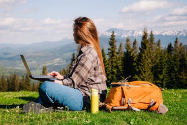 Ekose gömlek ve turuncu sırt çantalı bol saçlı genç, narin bir kız, yeşil çimlerin üzerinde, dağların arka planında oturuyor..