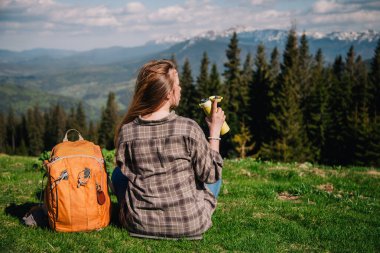 A young, slender girl with loose hair in a plaid shirt and jeans with an orange backpack drinks tea from a thermal mug sitting on the green grass against the backdrop of the mountains.