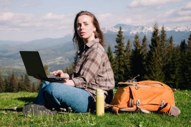 Ekose gömlek ve turuncu sırt çantalı bol saçlı genç, narin bir kız, yeşil çimlerin üzerinde, dağların arka planında oturuyor..
