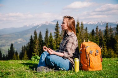 Ekose gömlekli, kot pantolonlu, turuncu sırt çantalı genç, narin bir kız yeşil çimlerin üzerinde, dağların arka planında meditasyon yapıyor..