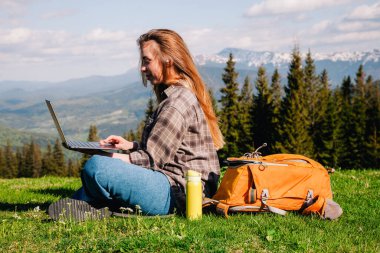 Ekose gömlek ve turuncu sırt çantalı bol saçlı genç, narin bir kız, yeşil çimlerin üzerinde, dağların arka planında oturuyor..