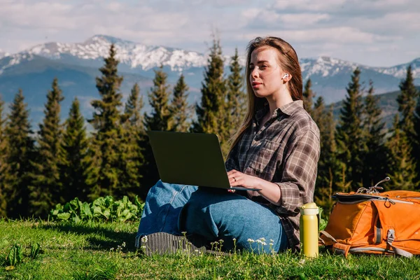 A young, slender girl with loose hair in a plaid shirt and jeans with an orange backpack works on a laptop sitting on green grass against the backdrop of mountains.