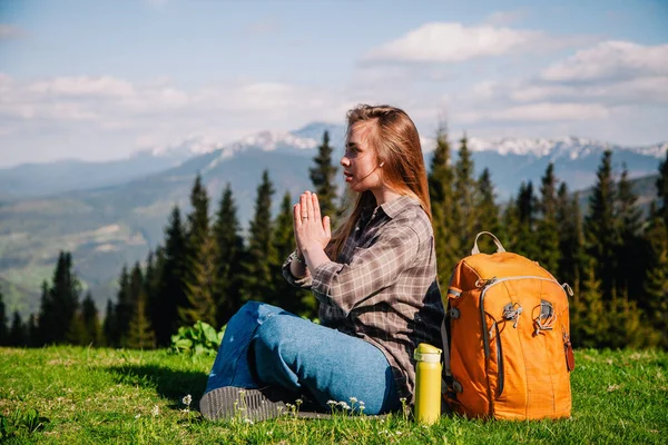 Ekose gömlekli, kot pantolonlu, turuncu sırt çantalı genç, narin bir kız yeşil çimlerin üzerinde, dağların arka planında meditasyon yapıyor..