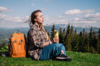 Genç, ince saçlı, ekose gömlekli, turuncu sırt çantalı, kot pantolonlu bir kız, dağların arka planında yeşil çimlerin üzerinde oturan termal bardaktan çay içer..