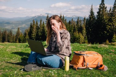 Ekose gömlek ve turuncu sırt çantalı bol saçlı genç, narin bir kız, yeşil çimlerin üzerinde, dağların arka planında oturuyor..