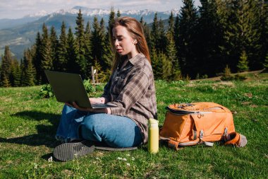 Ekose gömlek ve turuncu sırt çantalı bol saçlı genç, narin bir kız, yeşil çimlerin üzerinde, dağların arka planında oturuyor..