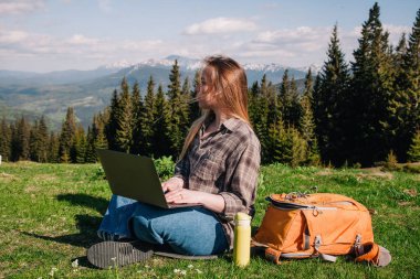 Ekose gömlek ve turuncu sırt çantalı bol saçlı genç, narin bir kız, yeşil çimlerin üzerinde, dağların arka planında oturuyor..