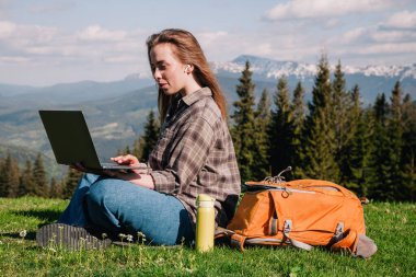 Ekose gömlek ve turuncu sırt çantalı bol saçlı genç, narin bir kız, yeşil çimlerin üzerinde, dağların arka planında oturuyor..