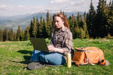 Ekose gömlek ve turuncu sırt çantalı bol saçlı genç, narin bir kız, yeşil çimlerin üzerinde, dağların arka planında oturuyor..