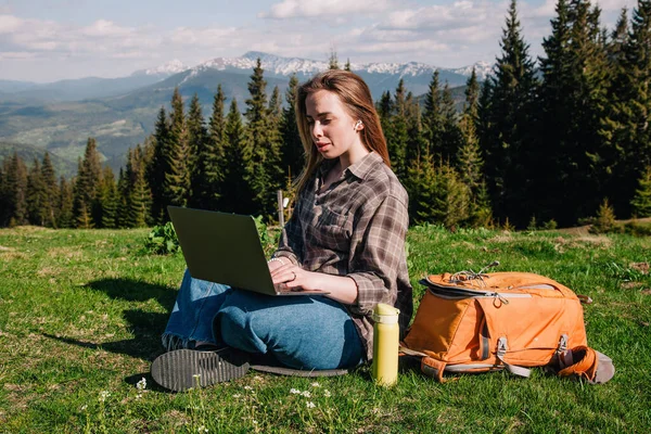 Ekose gömlek ve turuncu sırt çantalı bol saçlı genç, narin bir kız, yeşil çimlerin üzerinde, dağların arka planında oturuyor..