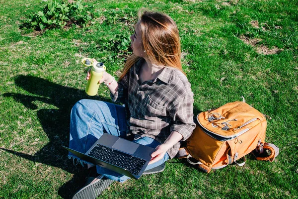 Молода струнка дівчина з розпущеним волоссям у плетеній сорочці та джинсах з помаранчевим рюкзаком працює на ноутбуці, сидячи на зеленій траві на тлі гір. вид зверху Стокове Зображення
