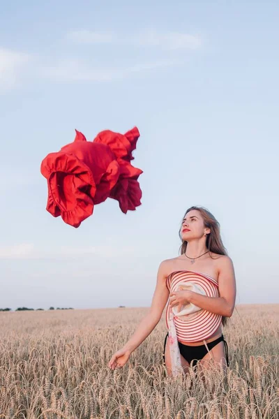 A young, slender girl with loose hair in a field of wheat hides behind a hat and tosses a red dress. High quality photo