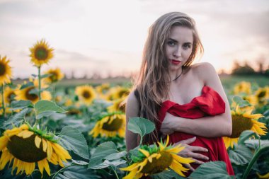 Genç, ince, gevşek saçlı bir kız, ayçiçeği tarlasında kırmızı bir elbiseyle kaplıdır. Yüksek kalite fotoğraf