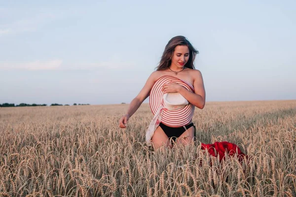 A young, slender girl with loose hair in a field of wheat hides behind a hat and tosses a red dress. High quality photo