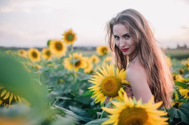 Genç, çıplak, ince saçlı bir kız gün batımında vücudunu ayçiçekleriyle kaplıyor. Yüksek kalite fotoğraf