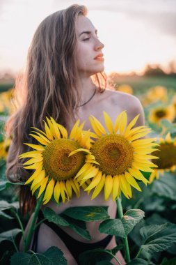 Genç, çıplak, ince saçlı bir kız gün batımında vücudunu ayçiçekleriyle kaplıyor. Yüksek kalite fotoğraf