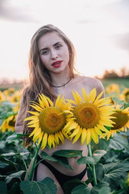 Genç, çıplak, ince saçlı bir kız gün batımında vücudunu ayçiçekleriyle kaplıyor. Yüksek kalite fotoğraf