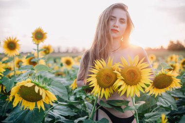 Genç, çıplak, ince saçlı bir kız gün batımında vücudunu ayçiçekleriyle kaplıyor. Yüksek kalite fotoğraf