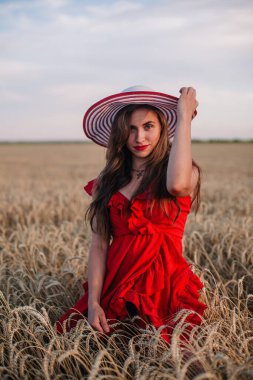 Genç, ince, parlak kırmızı elbiseli ve şapkalı seksi bir kız gün batımında buğday tarlasında duruyor. Yüksek kalite fotoğraf