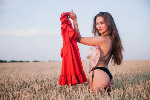 A young, slender, with loose hair in black panties in a field of wheat with a red dress in her hand. High quality photo