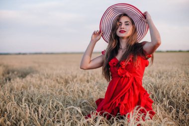 Genç, ince, parlak kırmızı elbiseli ve şapkalı seksi bir kız gün batımında buğday tarlasında duruyor. Yüksek kalite fotoğraf